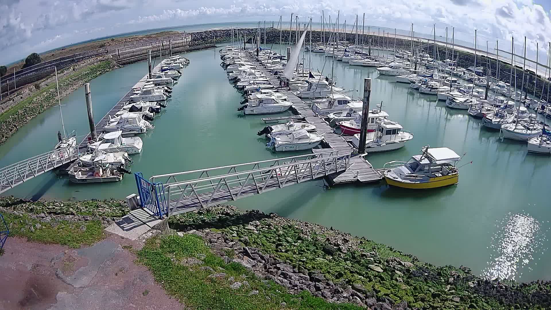 A marina filled with numerous boats on a partly cloudy day, with a metal walkway leading to a dock.