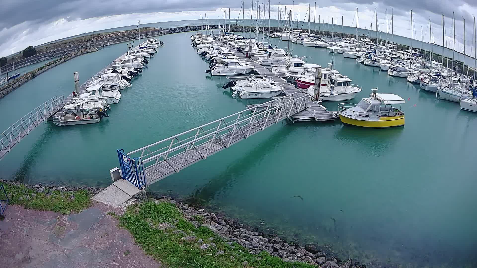 A marina filled with numerous boats of various sizes, under a cloudy sky, is connected by a metal walkway to the shore.
