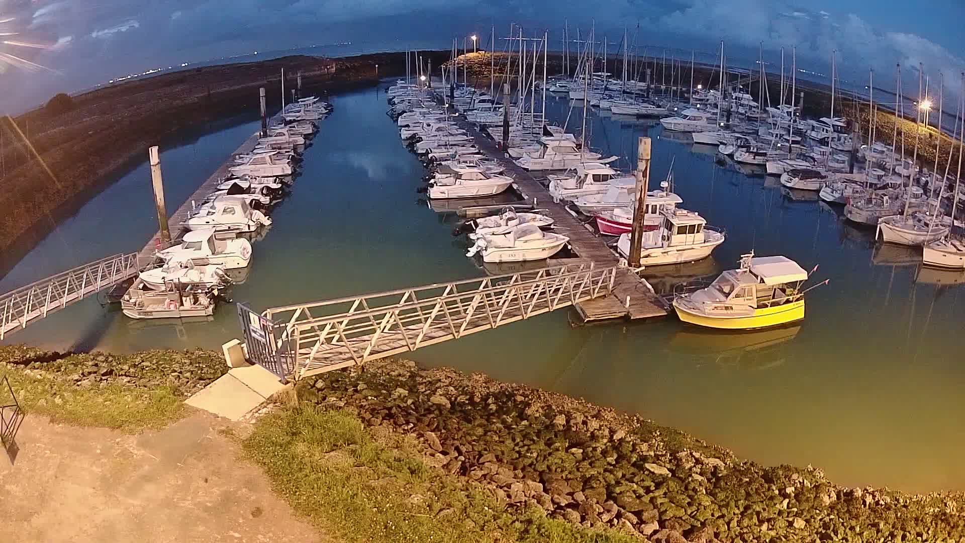 A marina at dusk filled with numerous boats docked along piers, with a metal walkway leading to the shore.