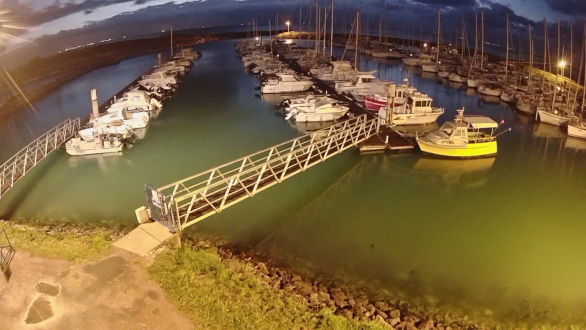 A marina at night, under a dark sky, shows many boats moored along docks connected by a metal walkway.
