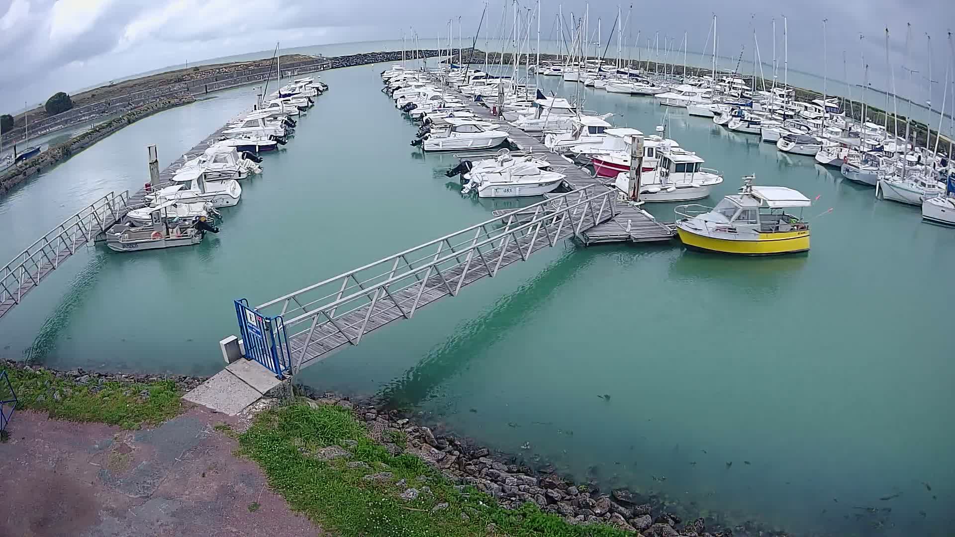 A marina filled with numerous boats and sailboats under a cloudy sky, connected by a metal footbridge.