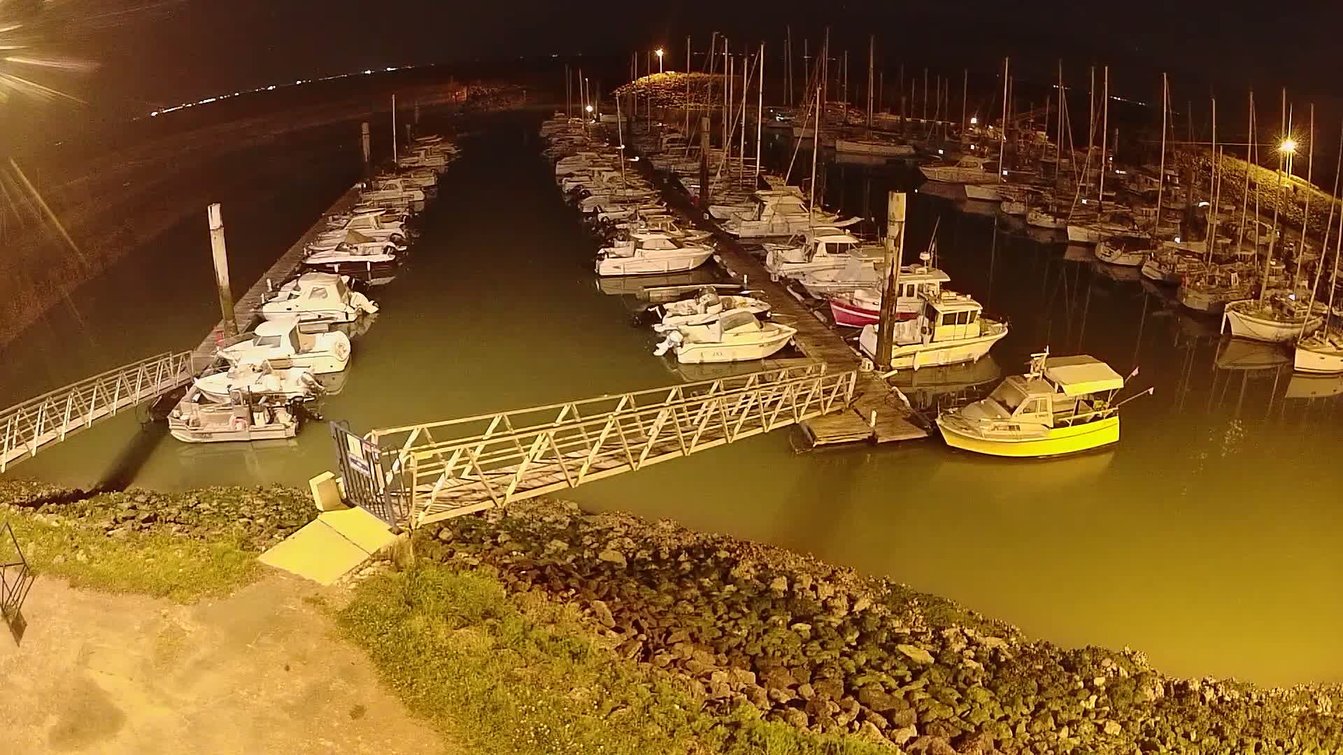 At night, a marina filled with numerous boats and yachts is illuminated, with a metal walkway extending from the shore to a dock.