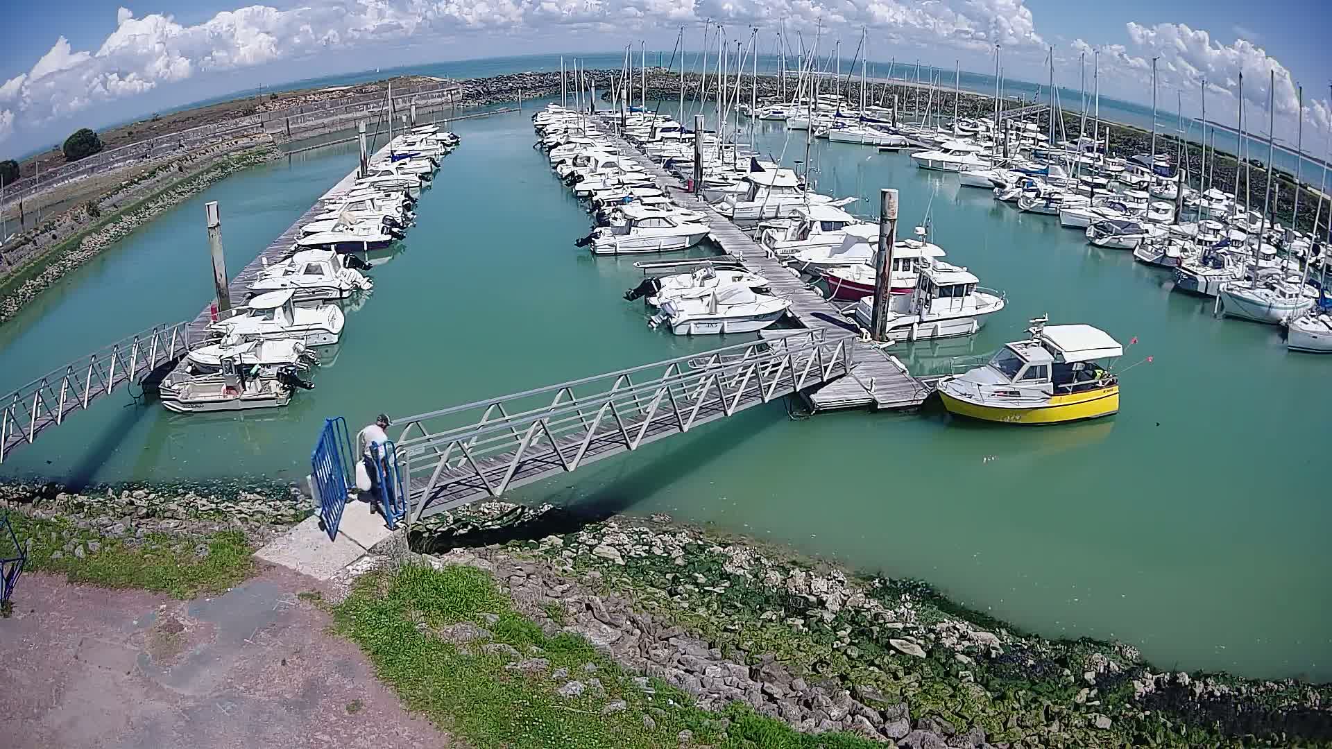 A marina filled with numerous boats and yachts under a partly cloudy sky, with a person walking across a metal footbridge to a dock.