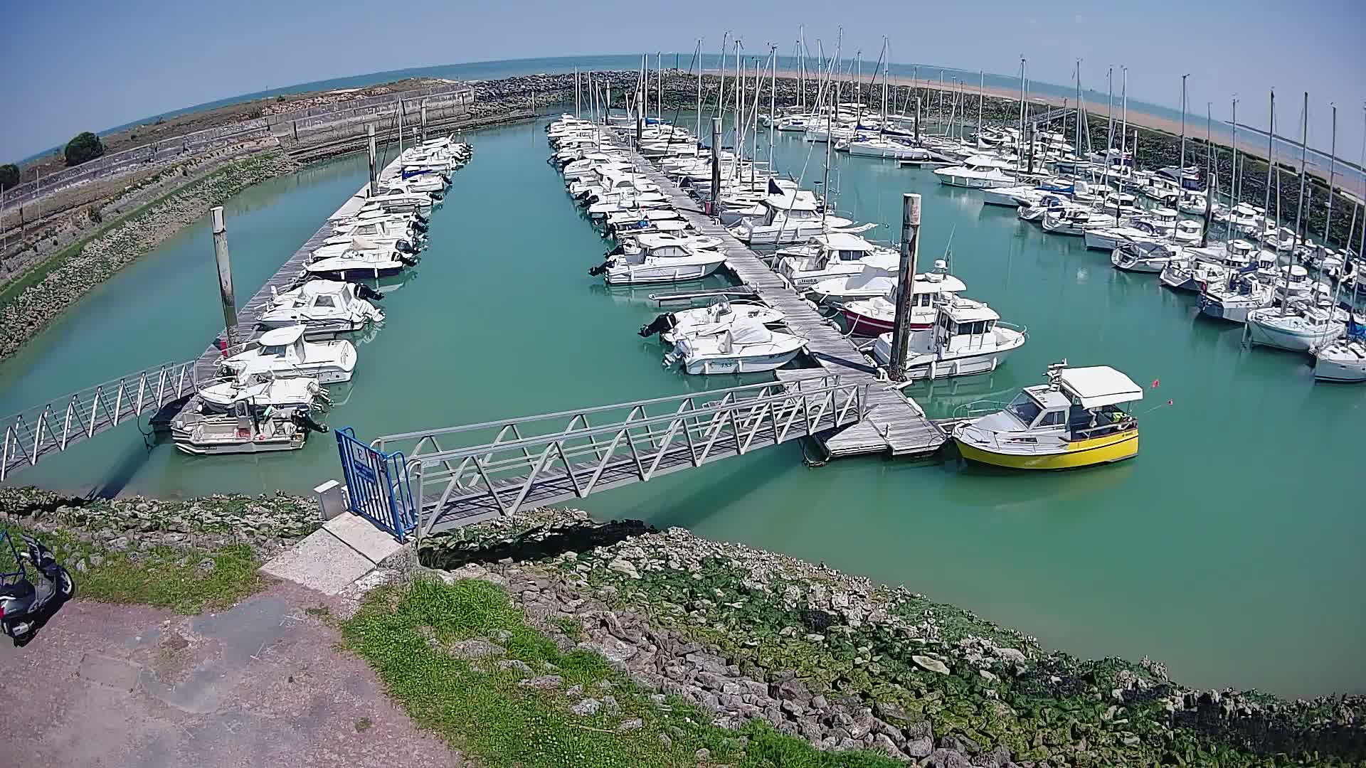 A marina filled with numerous boats of varying sizes is seen on a sunny day, with teal-colored water.