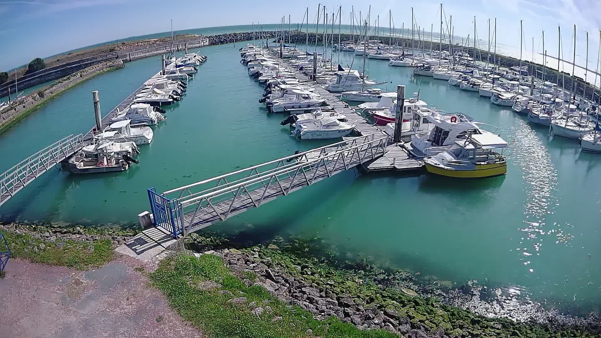A marina filled with numerous boats of varying sizes is shown on a sunny day, with calm, teal-colored water.