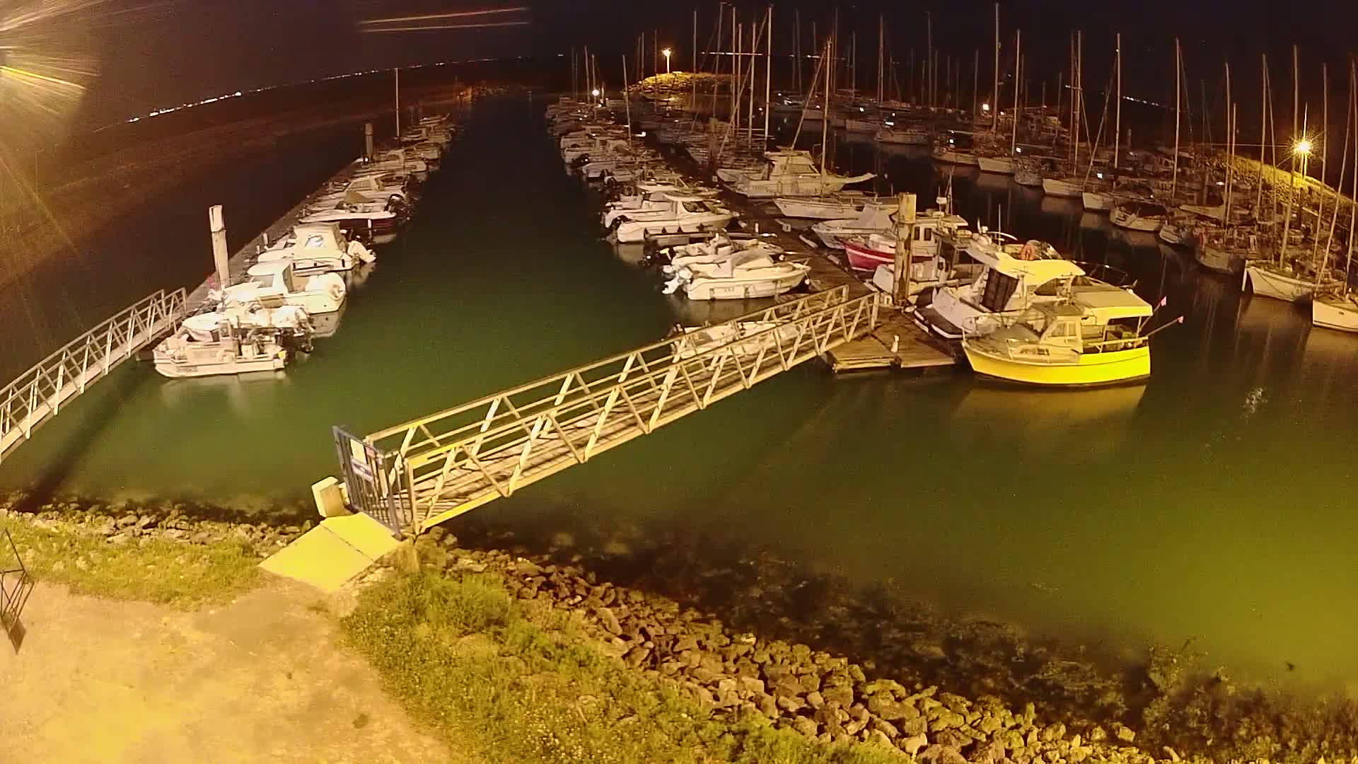 A marina at night, filled with numerous boats moored along docks, is connected by a metal walkway to the shore.