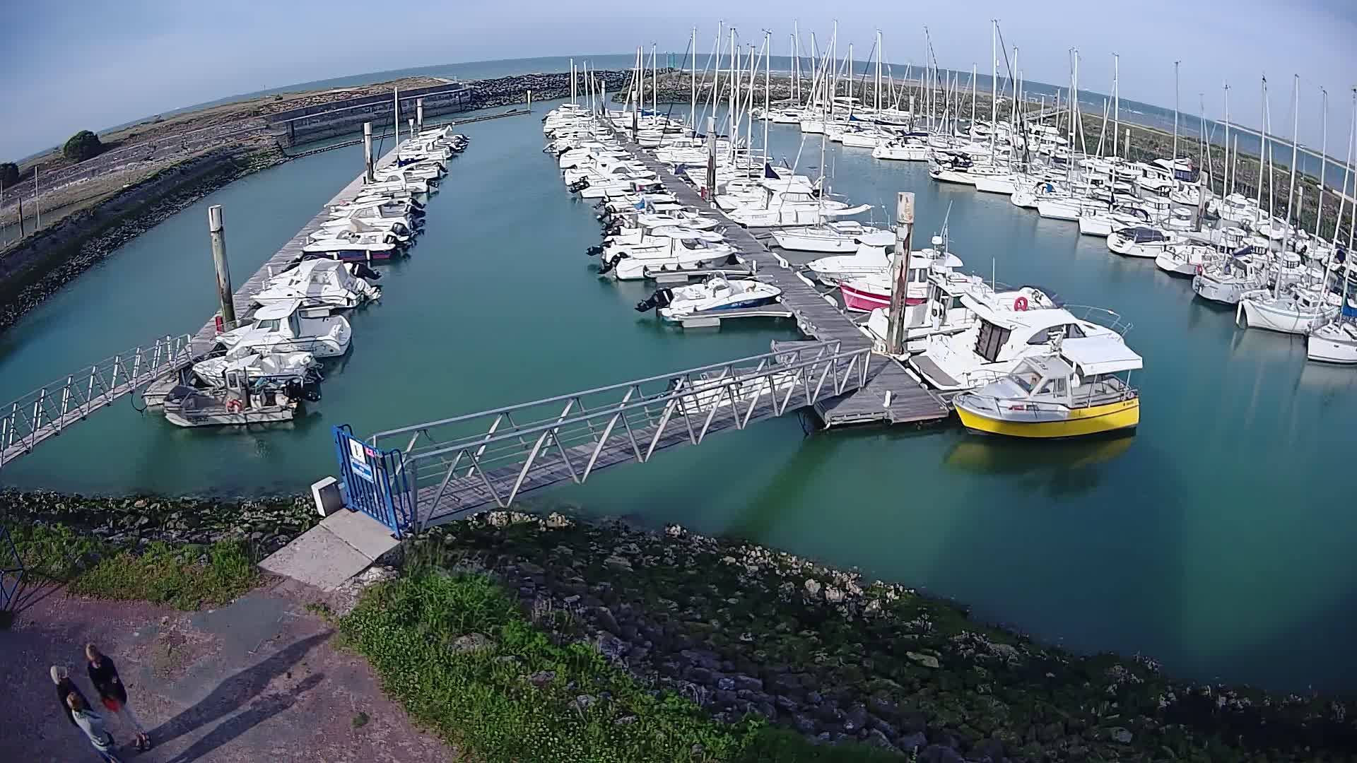 A marina filled with numerous sailboats and motorboats on a sunny day, connected by a metal walkway to the shore.