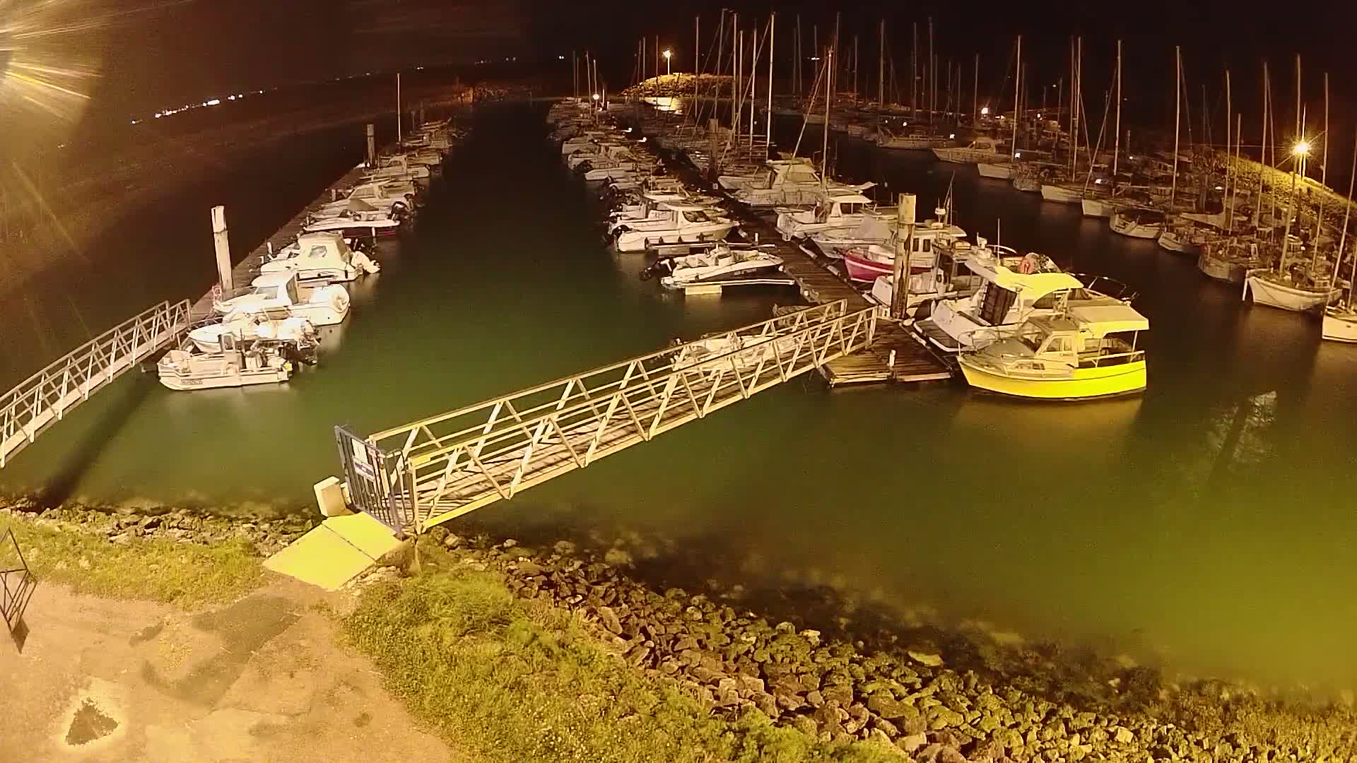 At night, a marina filled with docked boats is illuminated, with a metal walkway extending from the shore to a dock.