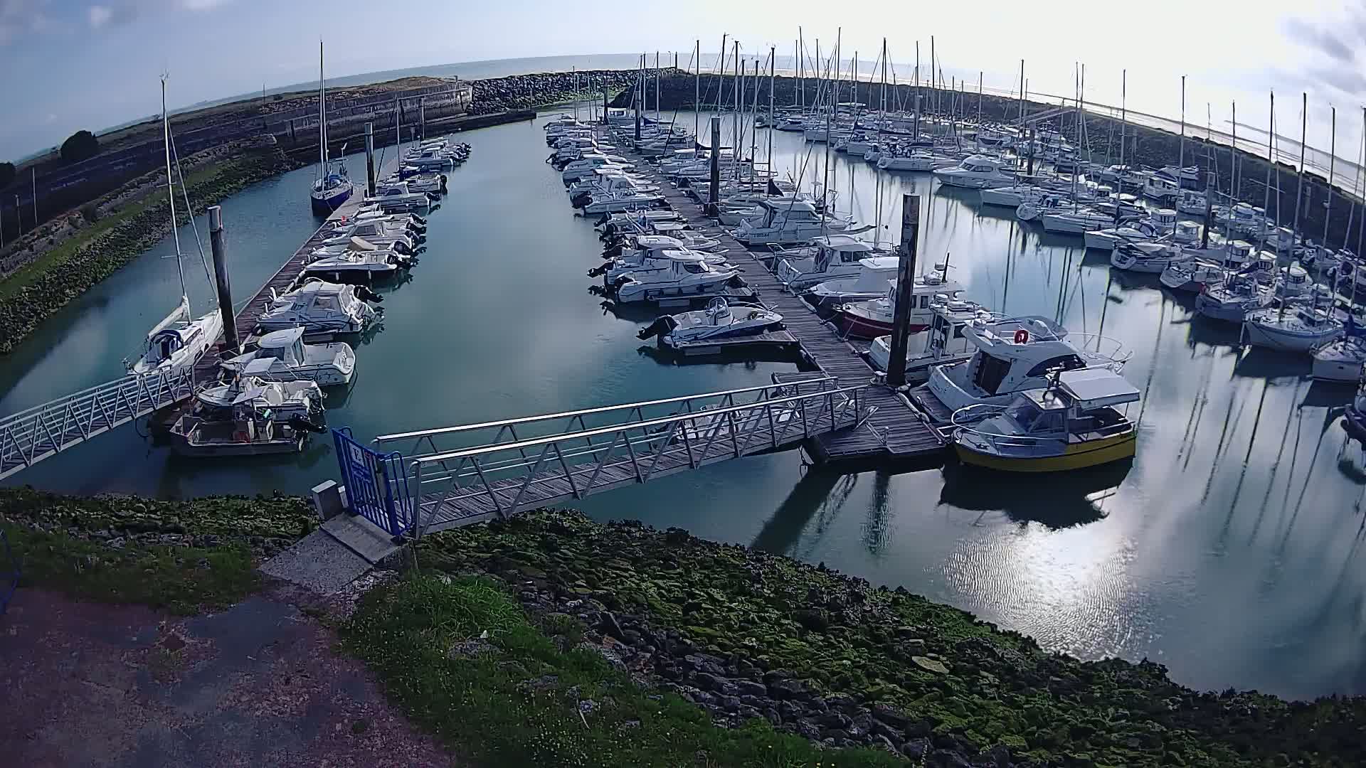 A marina filled with numerous boats of various sizes is shown on a sunny day, with a metal walkway leading to a dock.