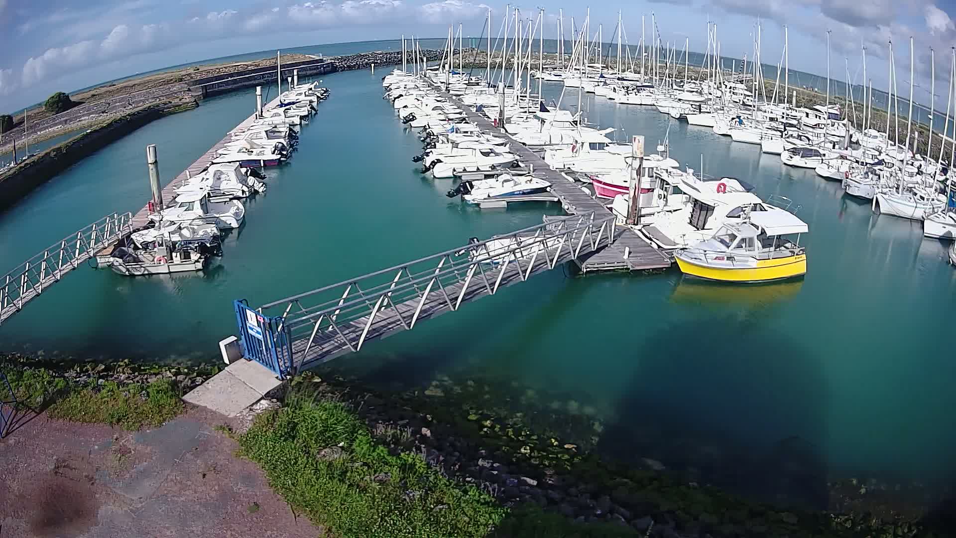 A marina filled with numerous sailboats and motorboats is seen on a sunny day with a partly cloudy sky.