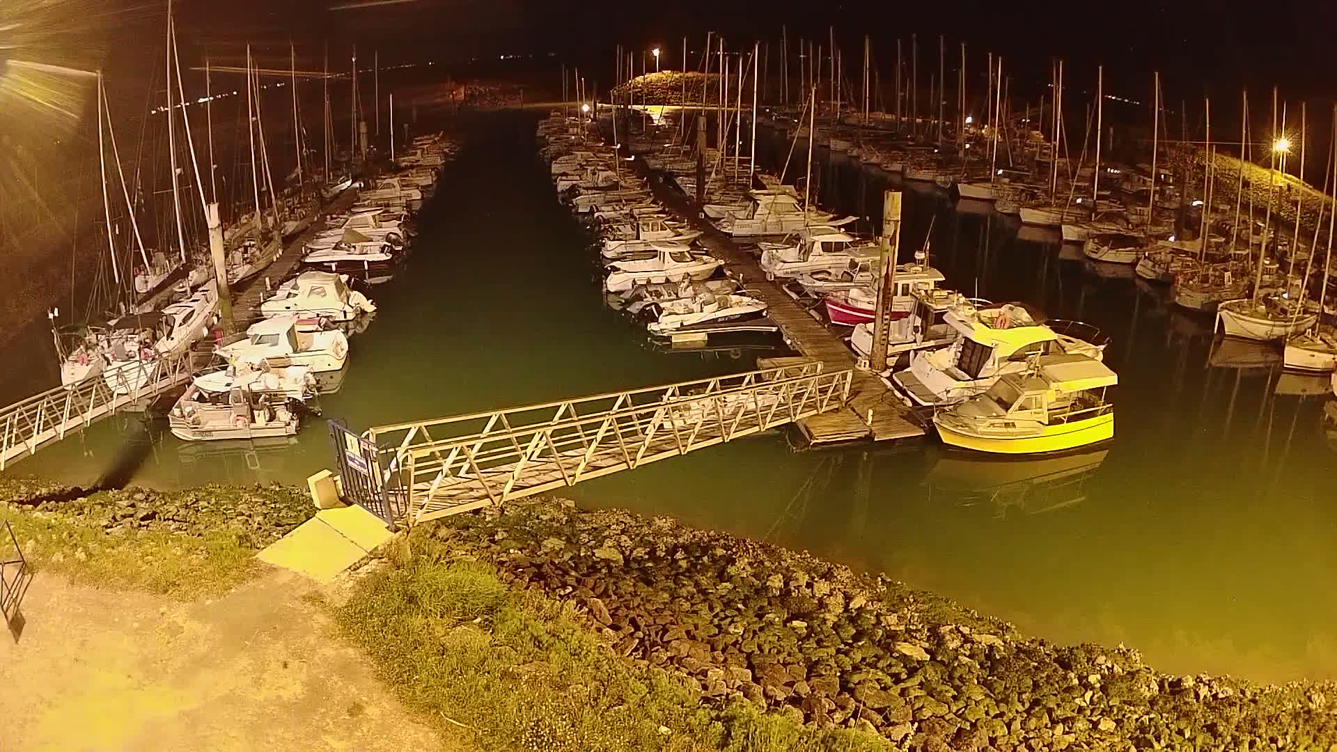 A marina at night, filled with numerous sailboats and motorboats docked along piers, illuminated by artificial lighting.