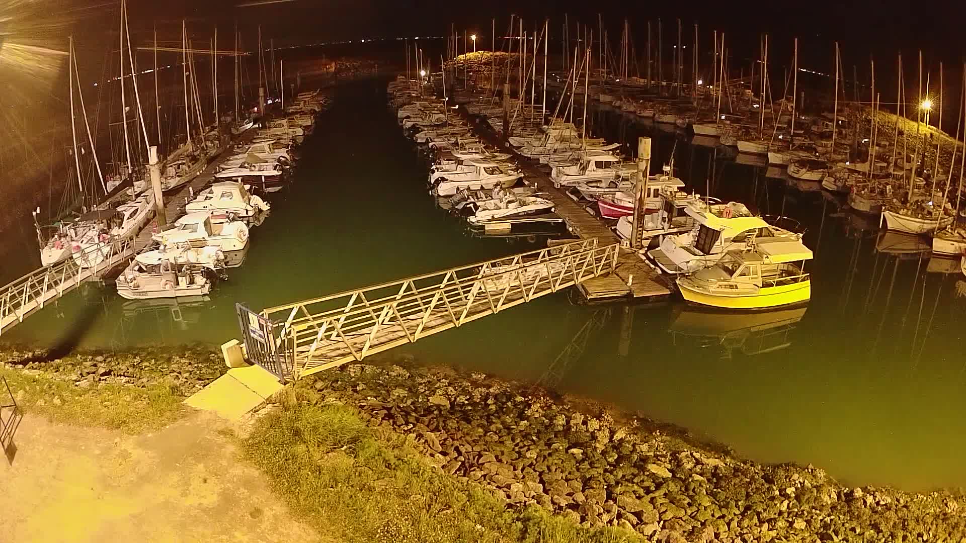 A marina filled with numerous boats at night, illuminated by artificial light, with a metal walkway connecting a dock to the shore.