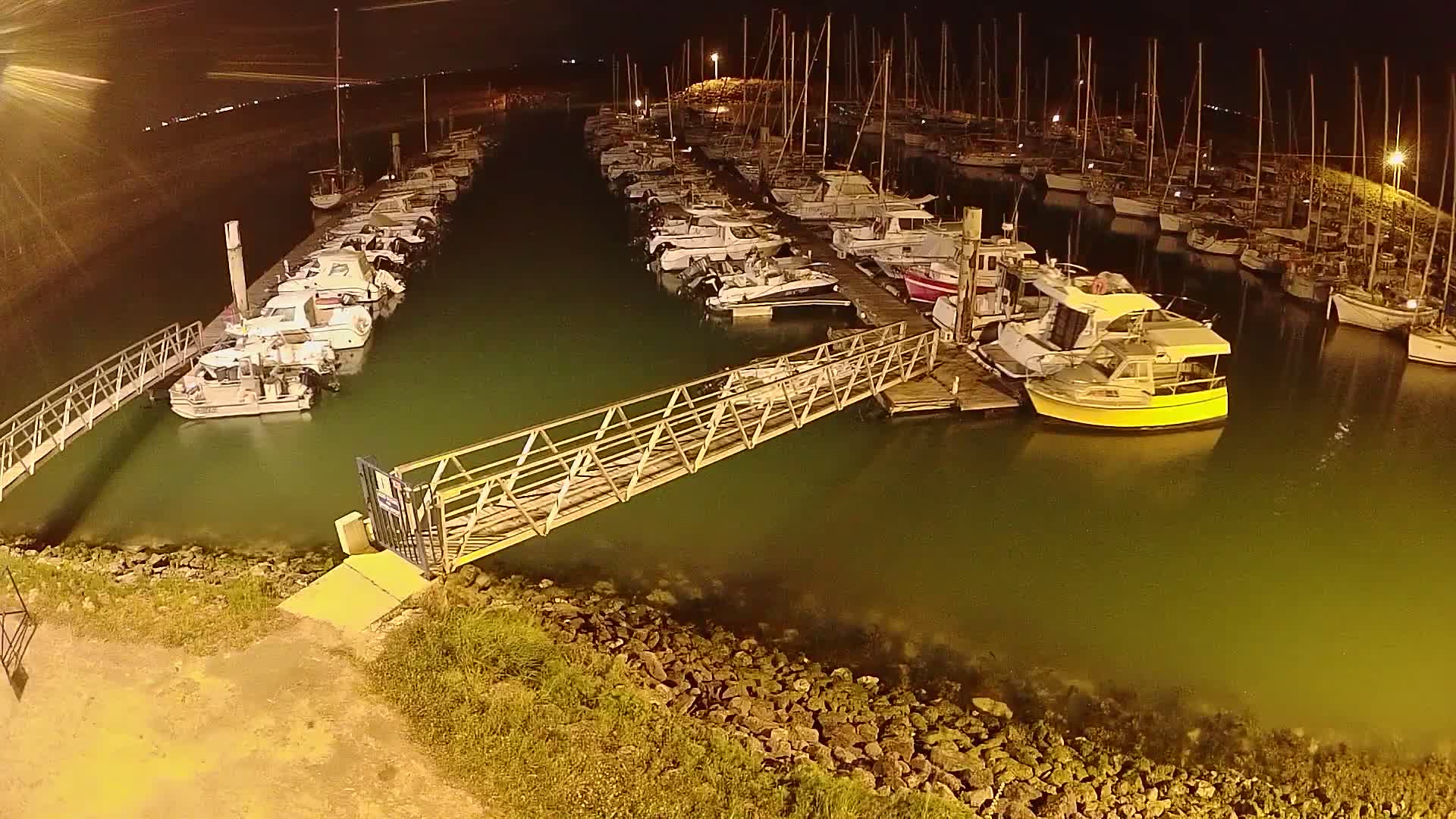 At night, a marina filled with numerous boats and yachts is illuminated, with a metal walkway connecting the docks.