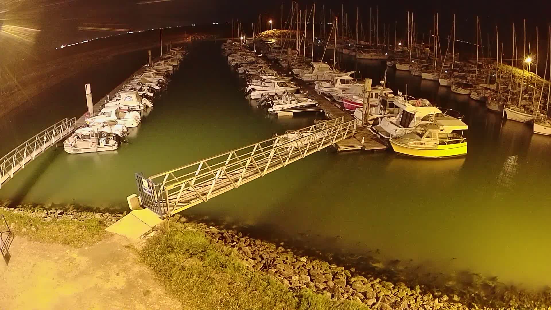 At night, numerous boats are docked at a marina, illuminated by artificial lights, with a metal walkway connecting the shore to a pier.