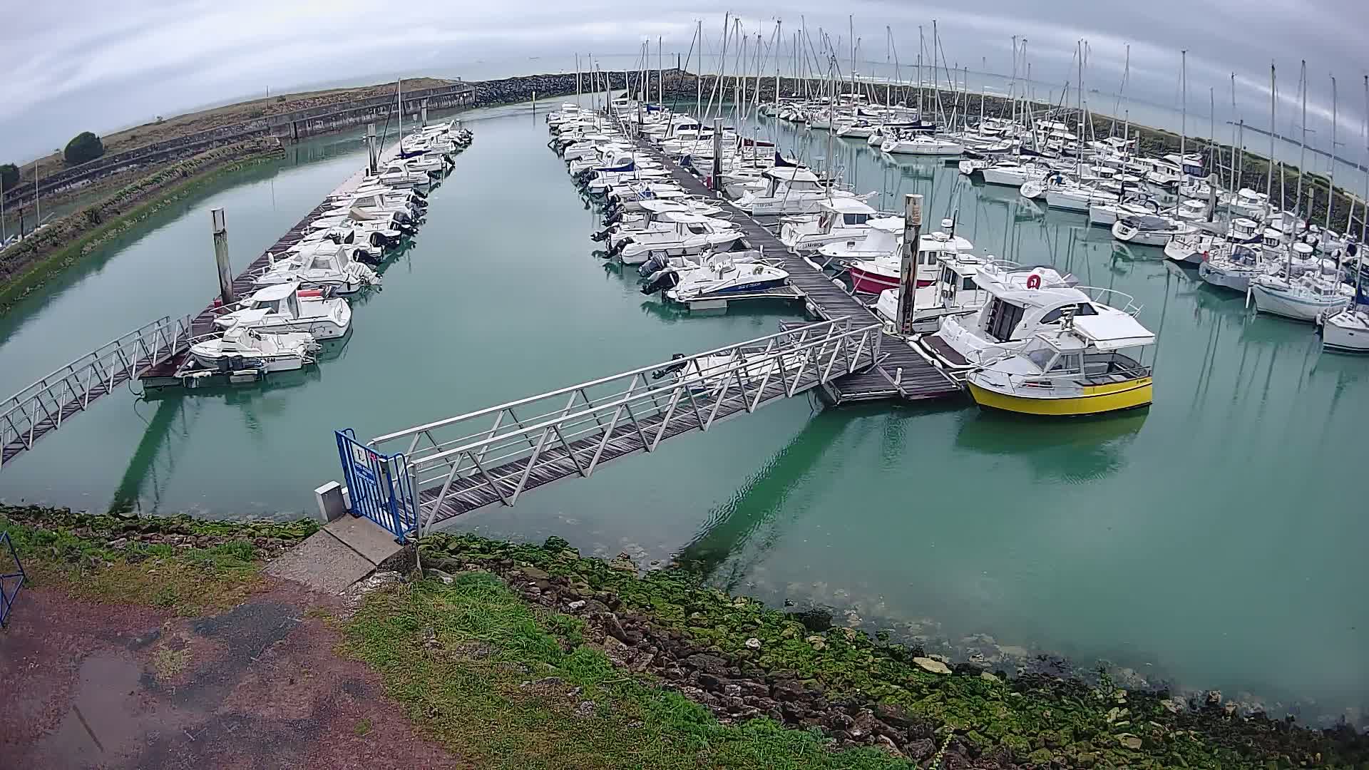 A marina with numerous boats docked along several piers under an overcast sky, connected by a metal walkway.