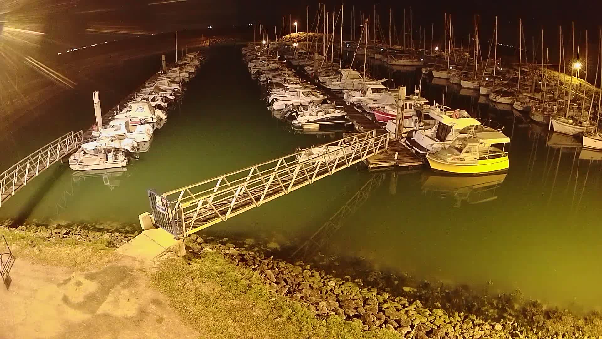 At night, many boats are docked at a marina with a metal walkway leading to a yellow boat.