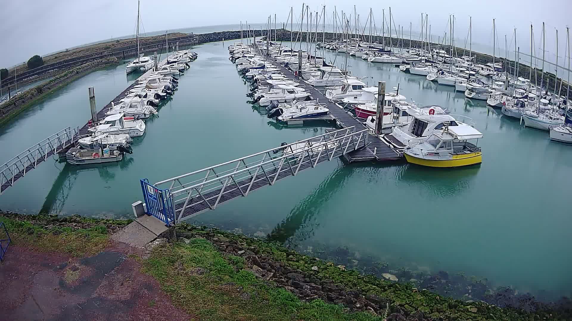 A marina filled with numerous boats on a cloudy day, with a metal walkway leading to a dock.
