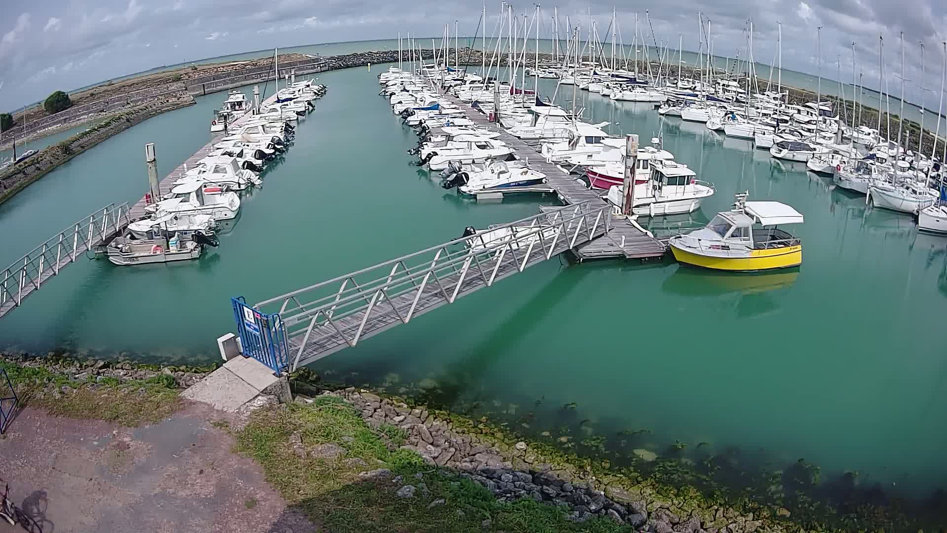 A marina filled with numerous sailboats and motorboats on a cloudy day, connected by a metal walkway to the shore.