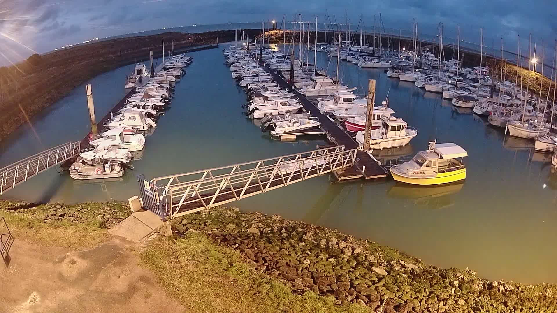 A marina at dusk filled with numerous docked sailboats and motorboats, connected by a metal walkway.