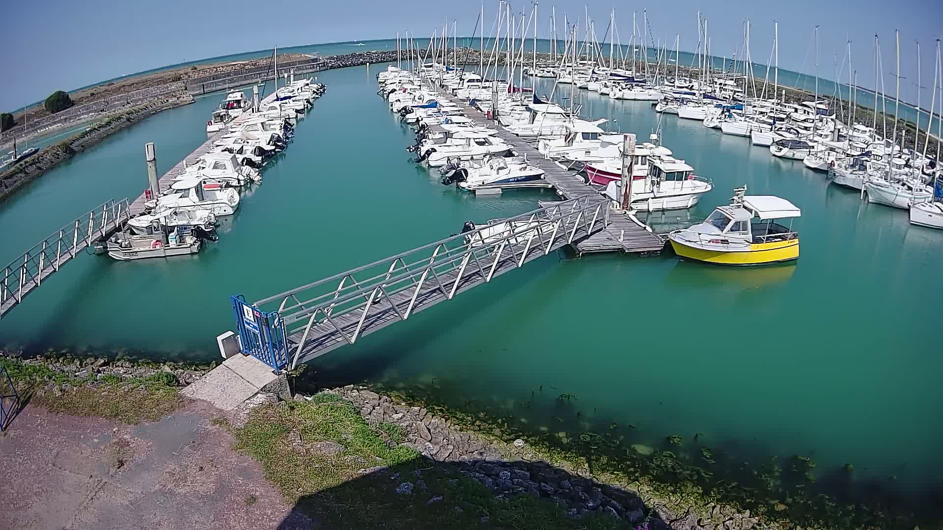 A marina with numerous boats docked along piers, connected by a metal walkway, under a clear sunny sky.