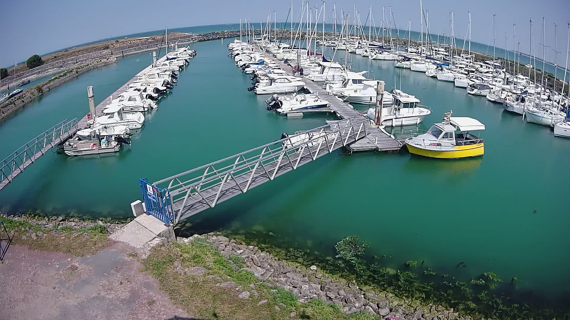 A marina filled with numerous boats of varying sizes under a clear, sunny sky, connected by a metal footbridge over teal water.