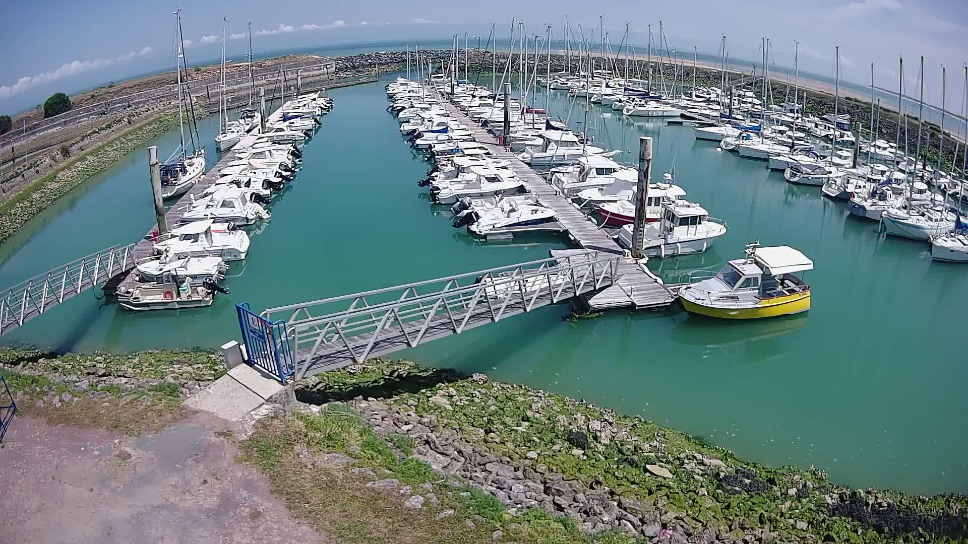 A marina filled with numerous boats and sailboats on a sunny day, connected by a metal walkway to the shore.
