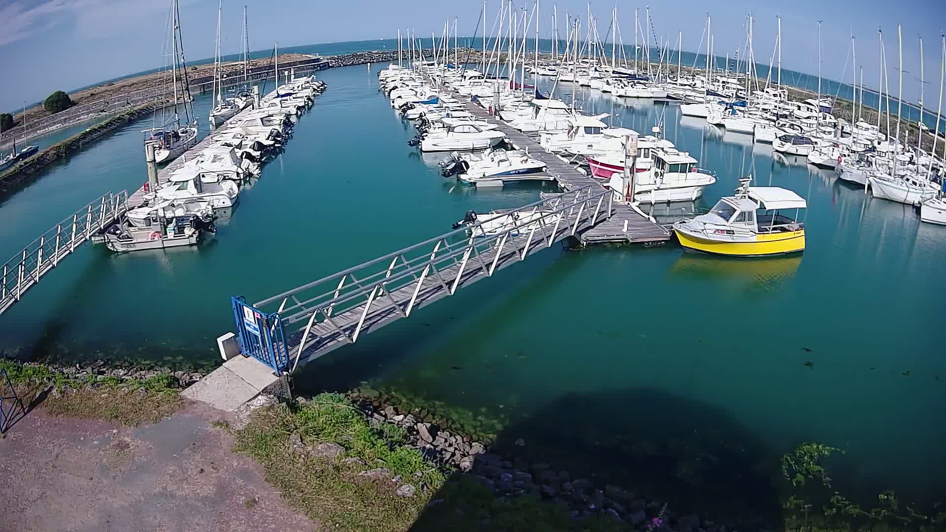 A marina filled with numerous boats of varying sizes, a small metallic footbridge connecting two docks, and clear teal water under a sunny sky.