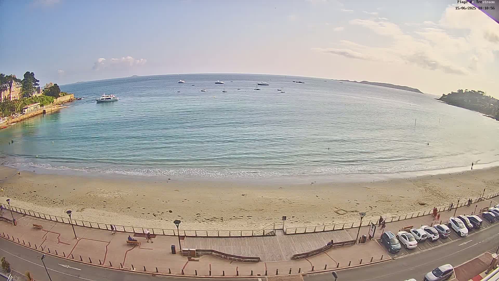 A sandy beach with a boardwalk and parked cars overlooks a calm, light blue ocean with several boats under a mostly clear sky.