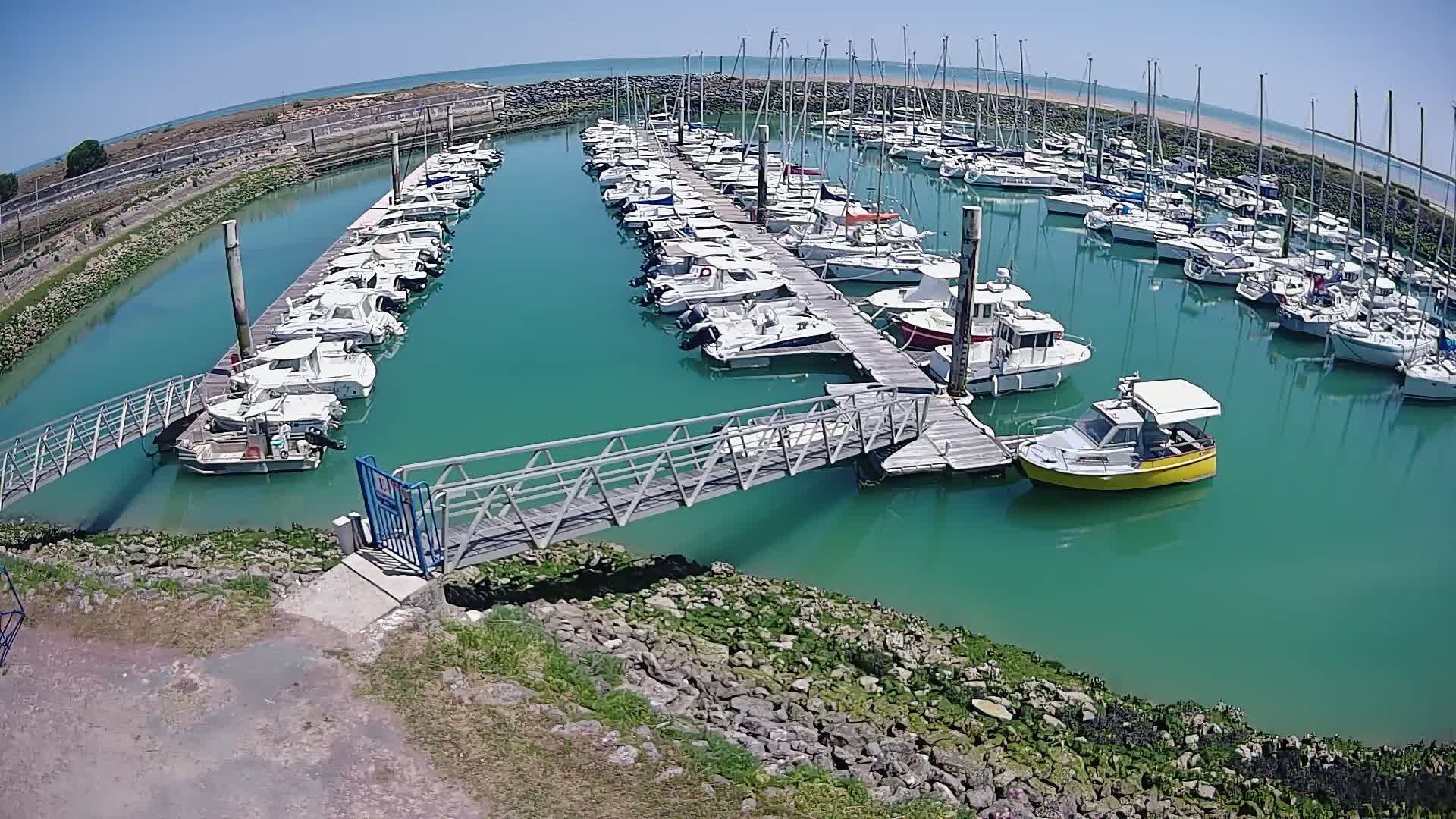 A marina filled with numerous boats of various sizes is nestled in a calm, teal-colored body of water under a clear, sunny sky.