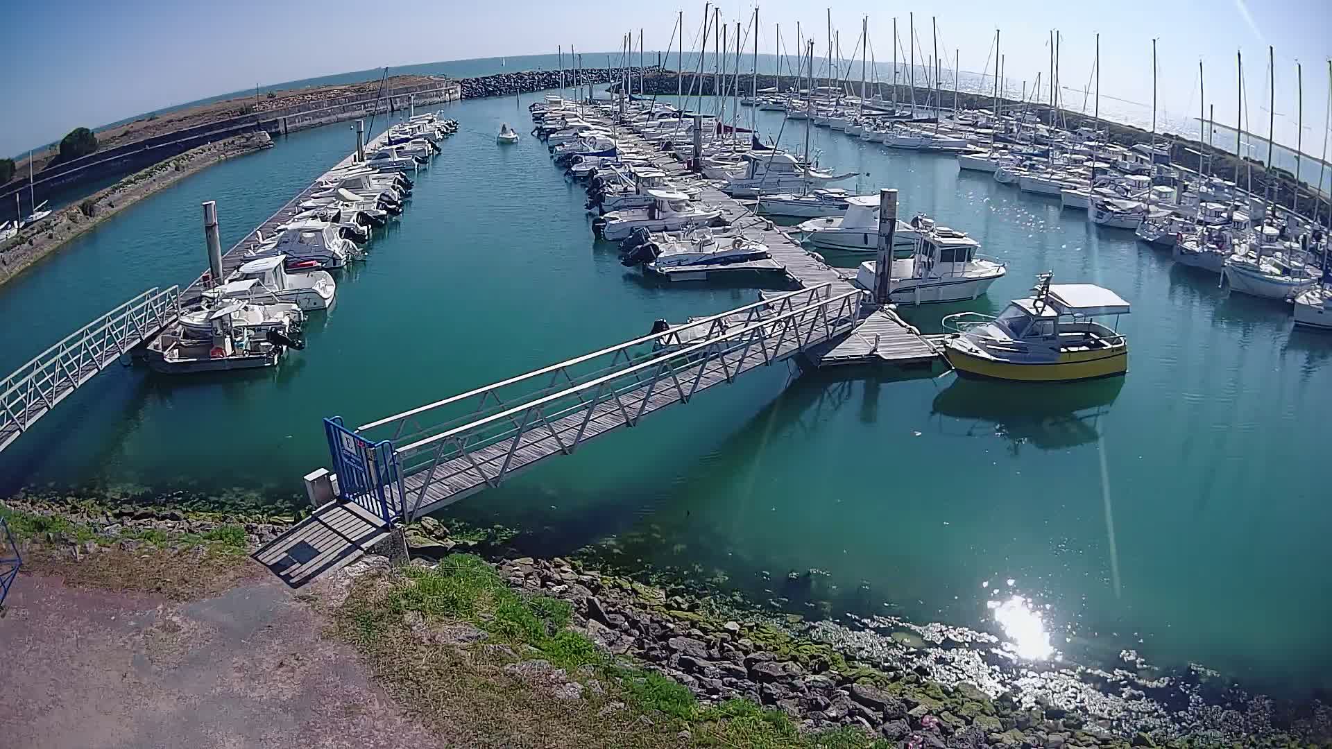 A marina filled with numerous boats on a sunny day, connected by a metal walkway over teal-colored water.