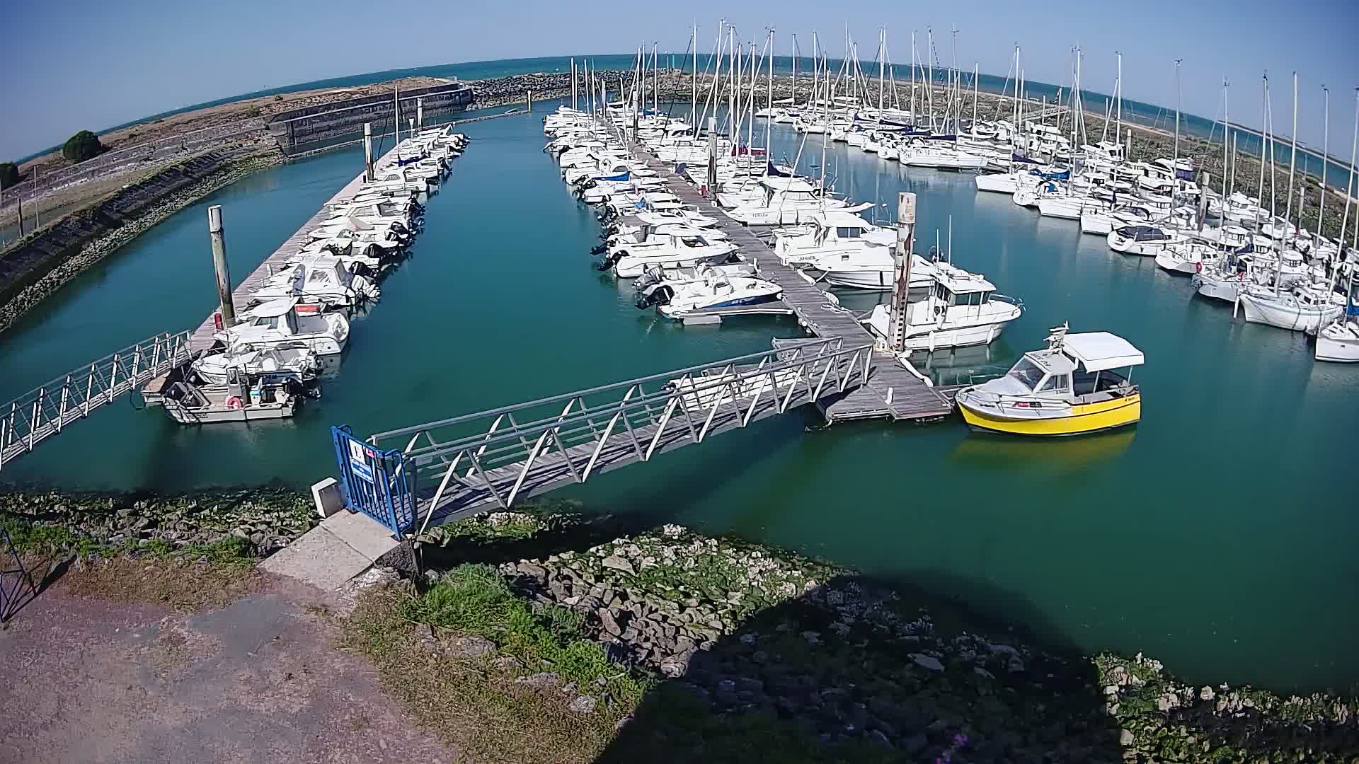 A marina filled with numerous boats of varying sizes on a sunny day, connected by a metal walkway to the shore.