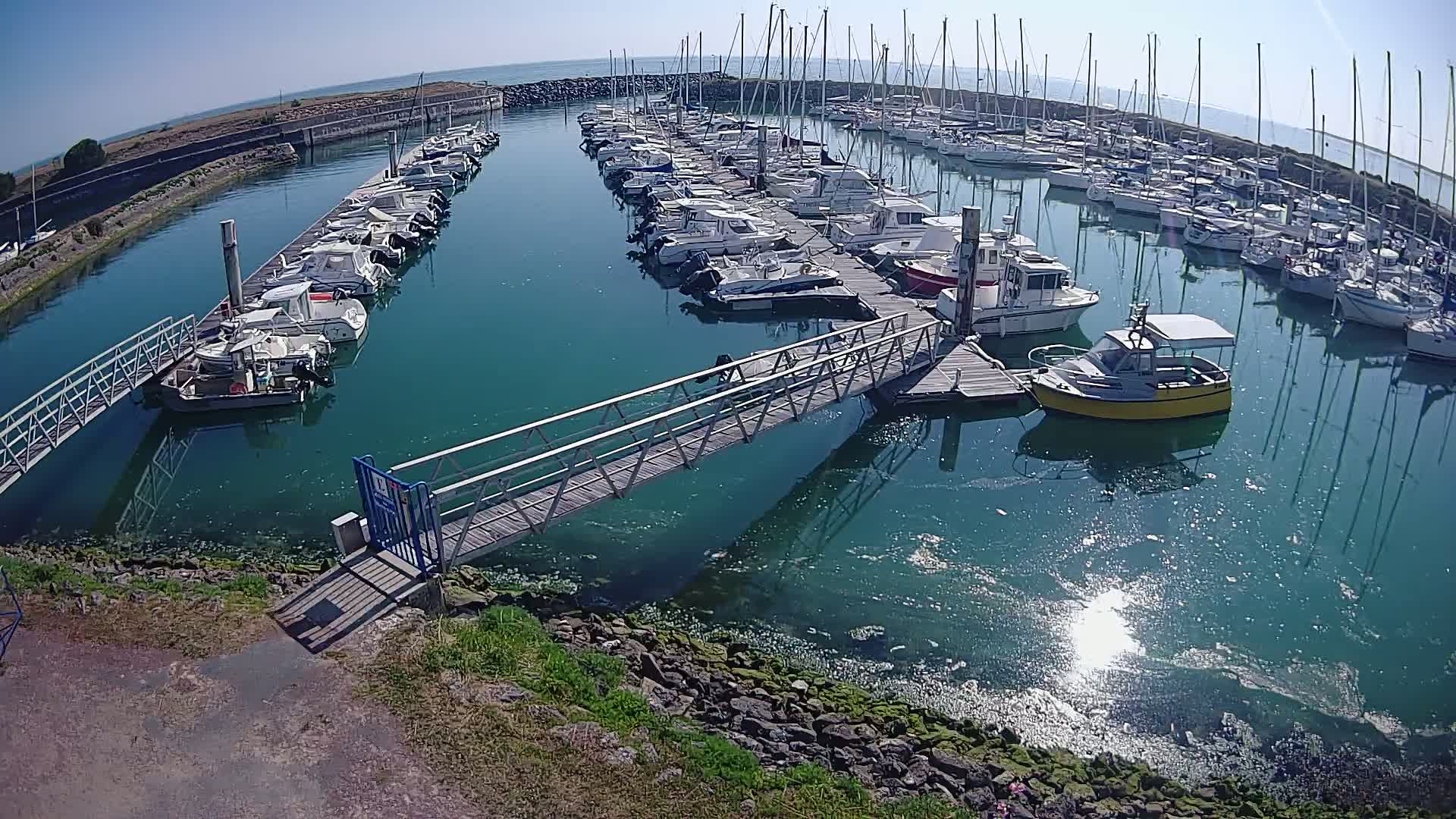 A marina filled with numerous sailboats and motorboats on a sunny day, with a metal walkway connecting a dock to the shore.
