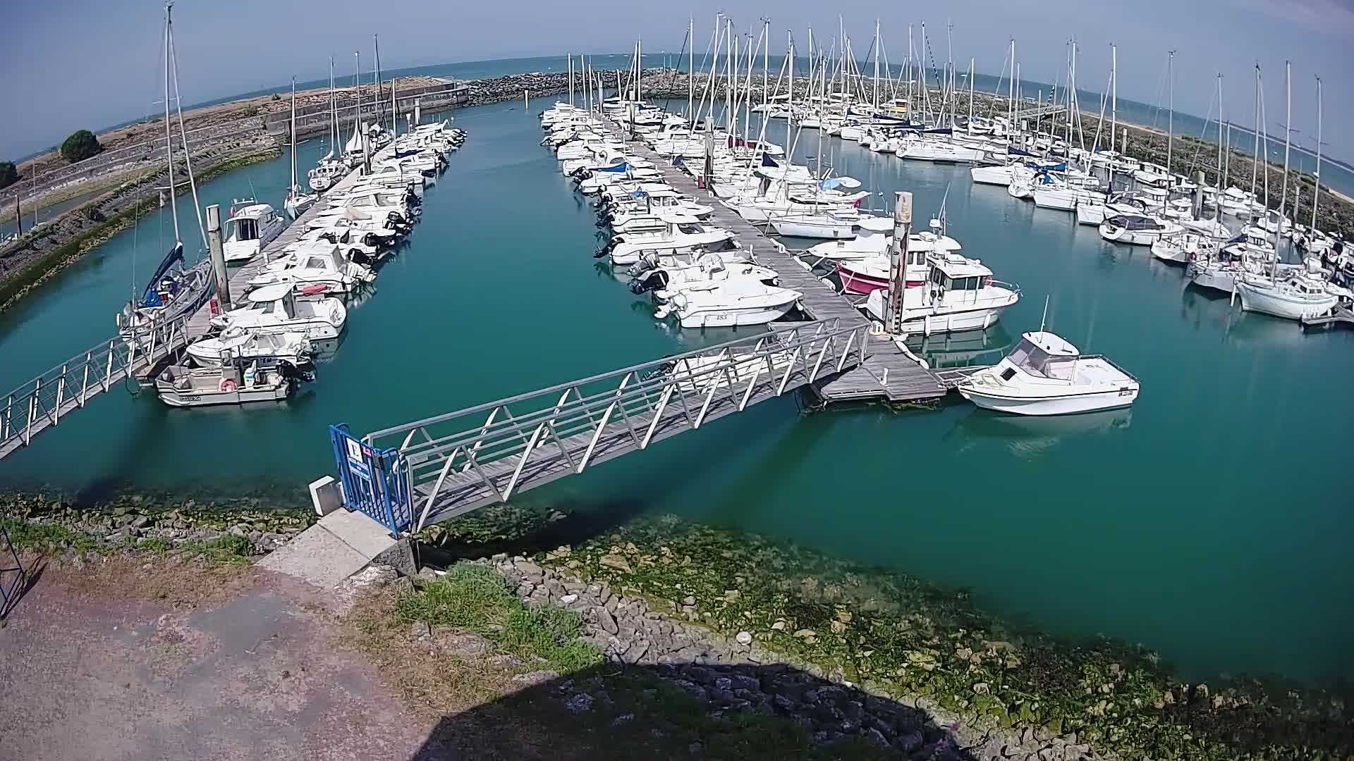 A marina filled with numerous sailboats and motorboats on a sunny day, connected by a metal walkway over teal water.