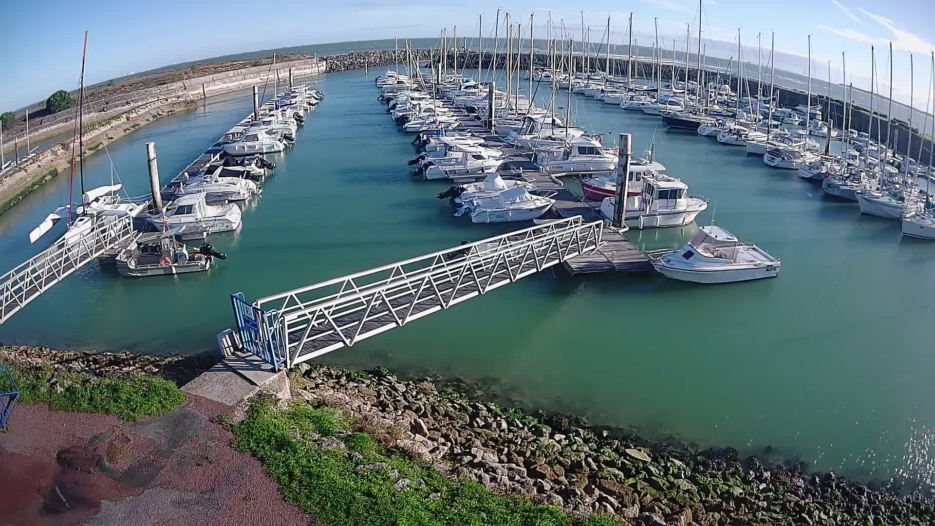 Numerous boats, including sailboats and motorboats, are docked in a bustling marina under a clear, sunny sky, with calm greenish-blue water reflecting the bright conditions.