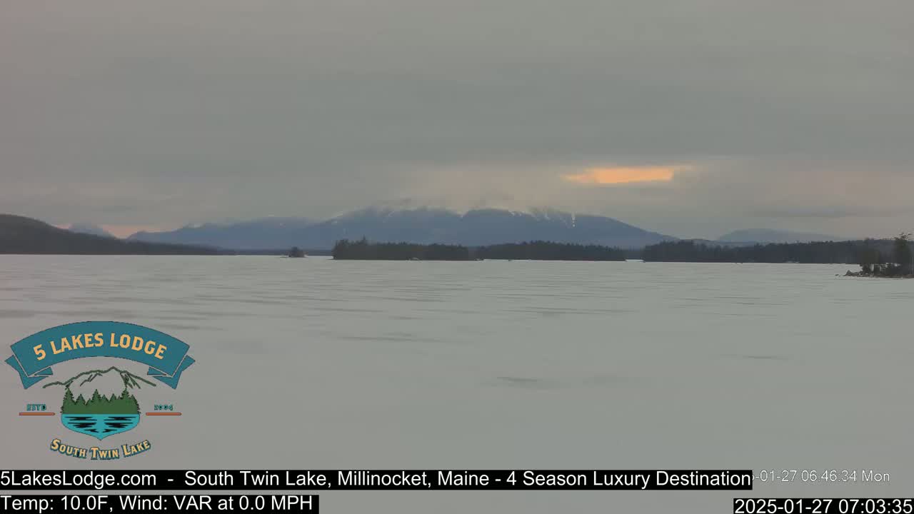 A frozen lake stretches to snow-covered mountains under a mostly cloudy sky.