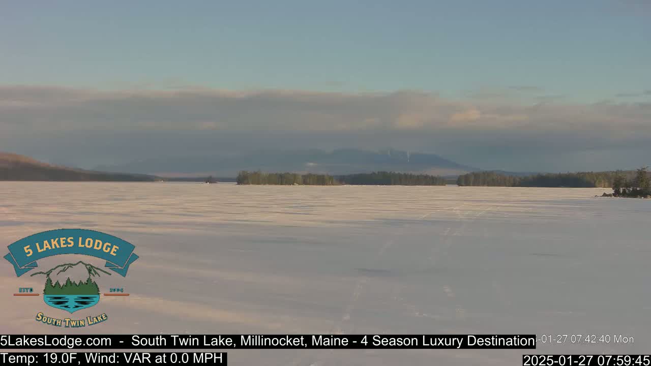 A frozen lake stretches to a distant, snow-covered mountain range under a partly cloudy sky.