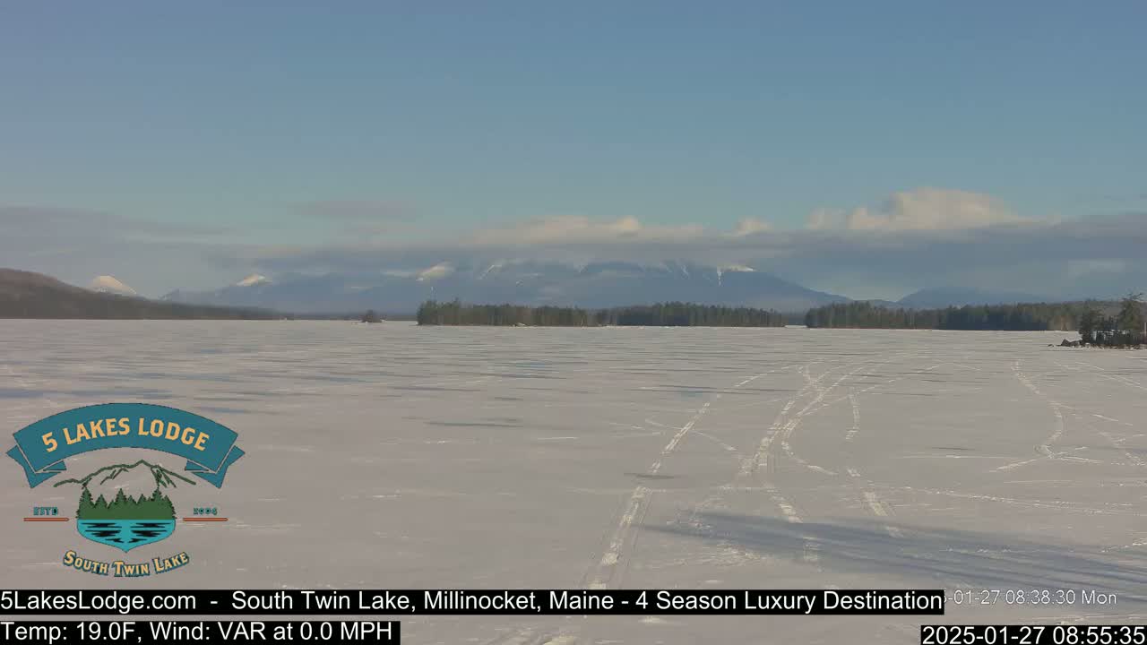 A frozen lake stretches to snow-covered mountains under a partly cloudy sky.