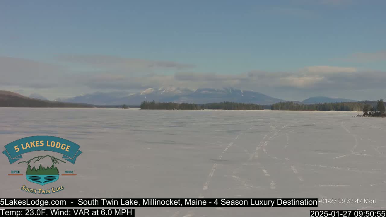 A frozen lake stretches toward snow-capped mountains under a partly cloudy sky.