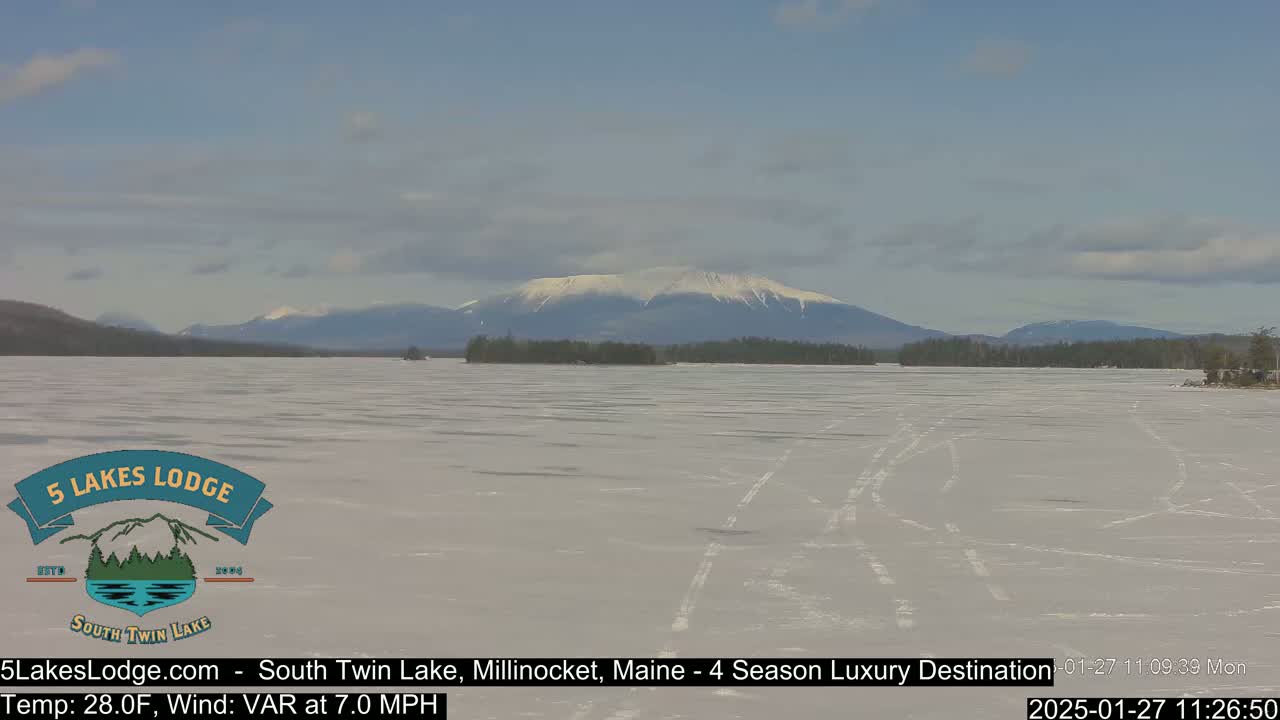 A frozen lake stretches towards snow-capped mountains under a partly cloudy sky.