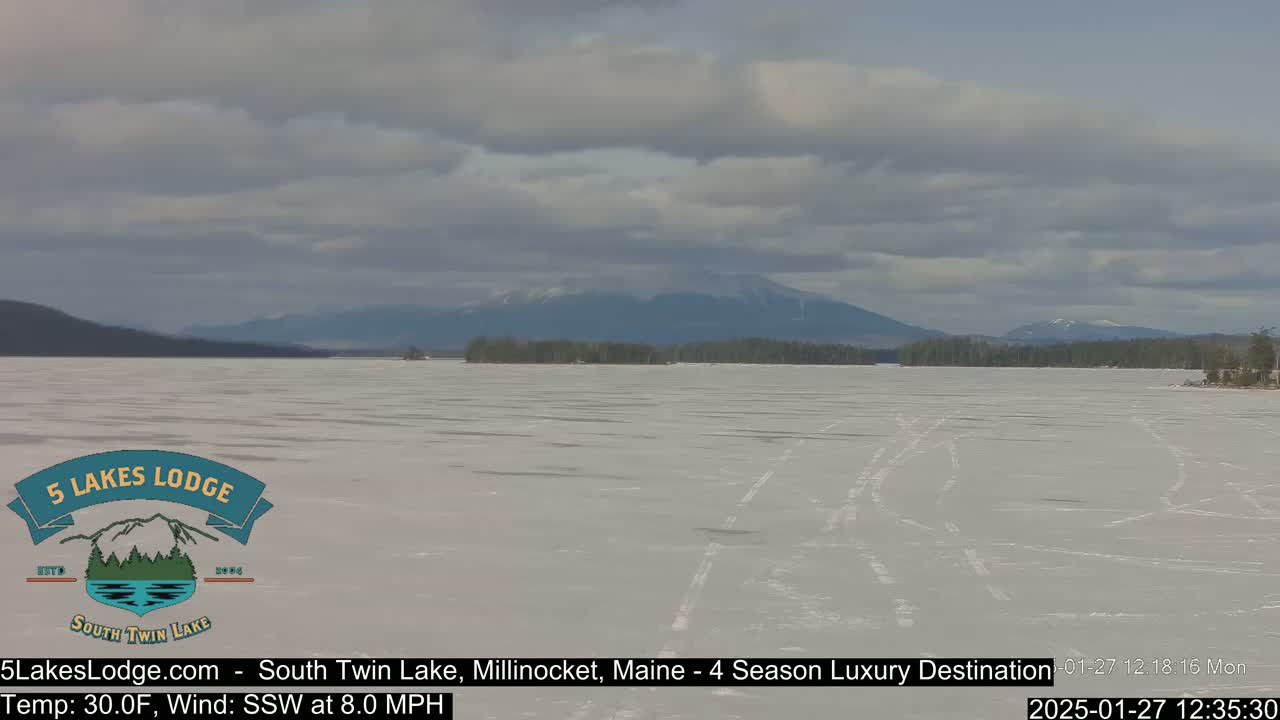A frozen lake stretches to snow-capped mountains under a cloudy sky.