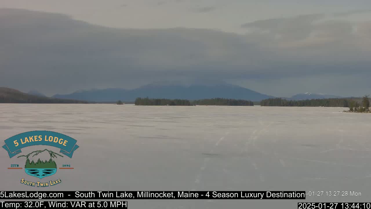 A frozen lake stretches to distant, snow-capped mountains under a cloudy sky.