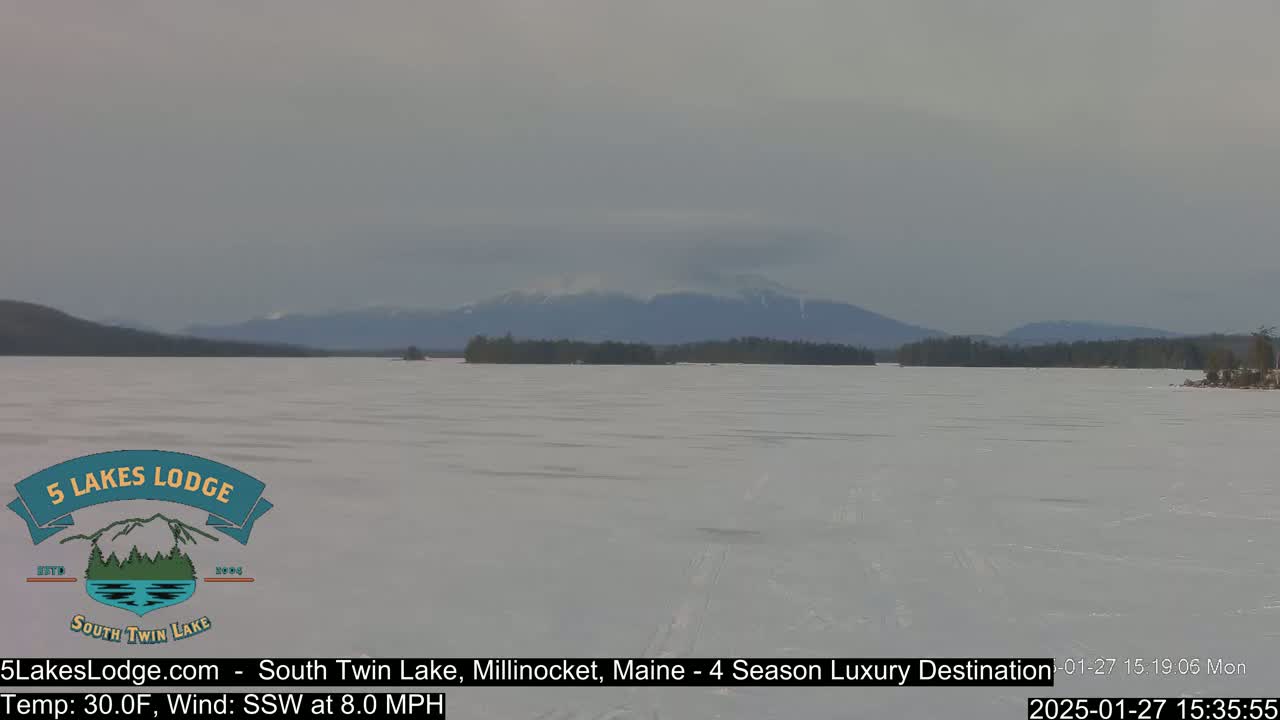 A frozen lake stretches toward distant mountains under an overcast sky, with a light south-southwesterly wind.