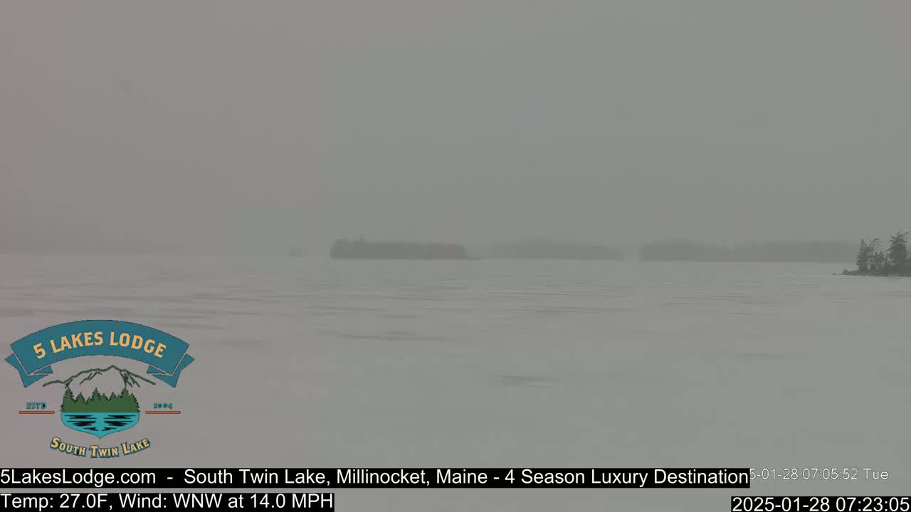 A hazy, snowy scene shows a calm lake with distant, indistinct landmasses under a gray sky.