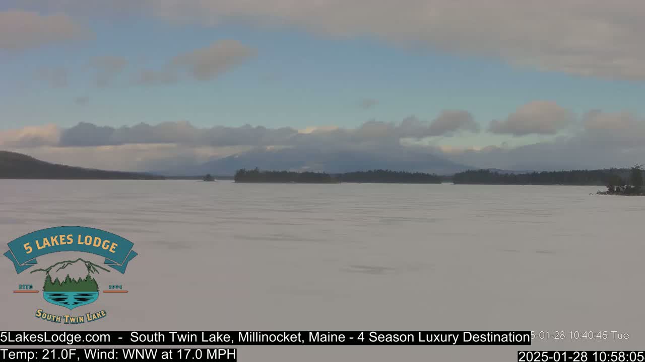 A snow-covered lake stretches to a distant, tree-lined shore under a partly cloudy sky.