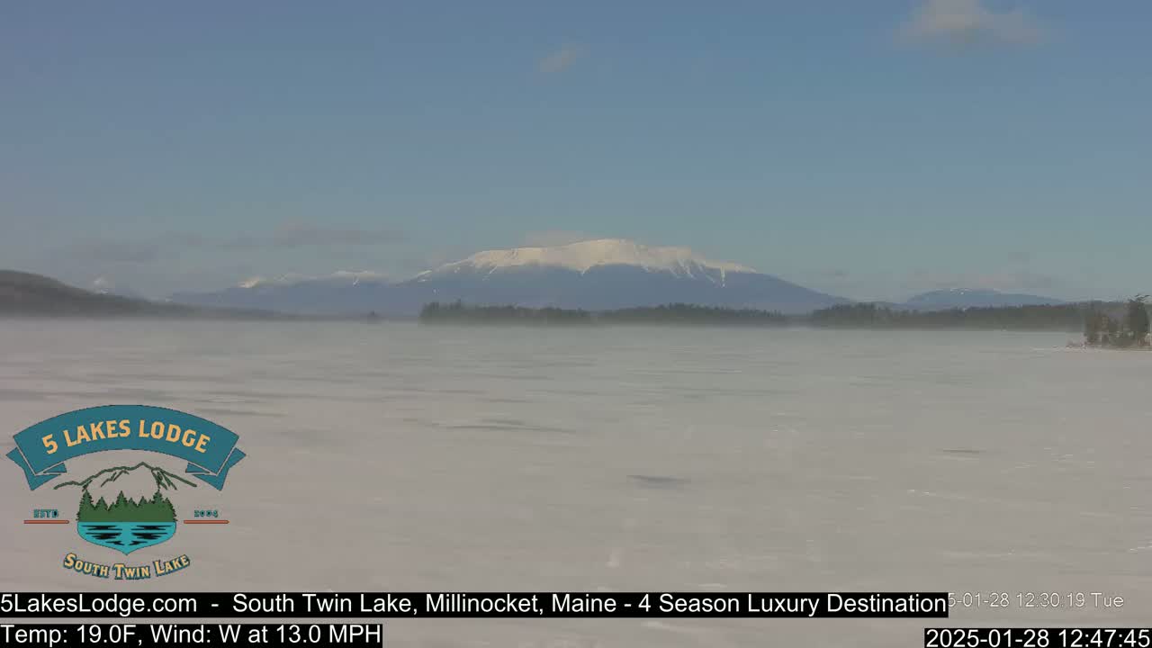 A partially frozen lake with a misty atmosphere shows a snow-capped mountain range in the background under a clear sky.