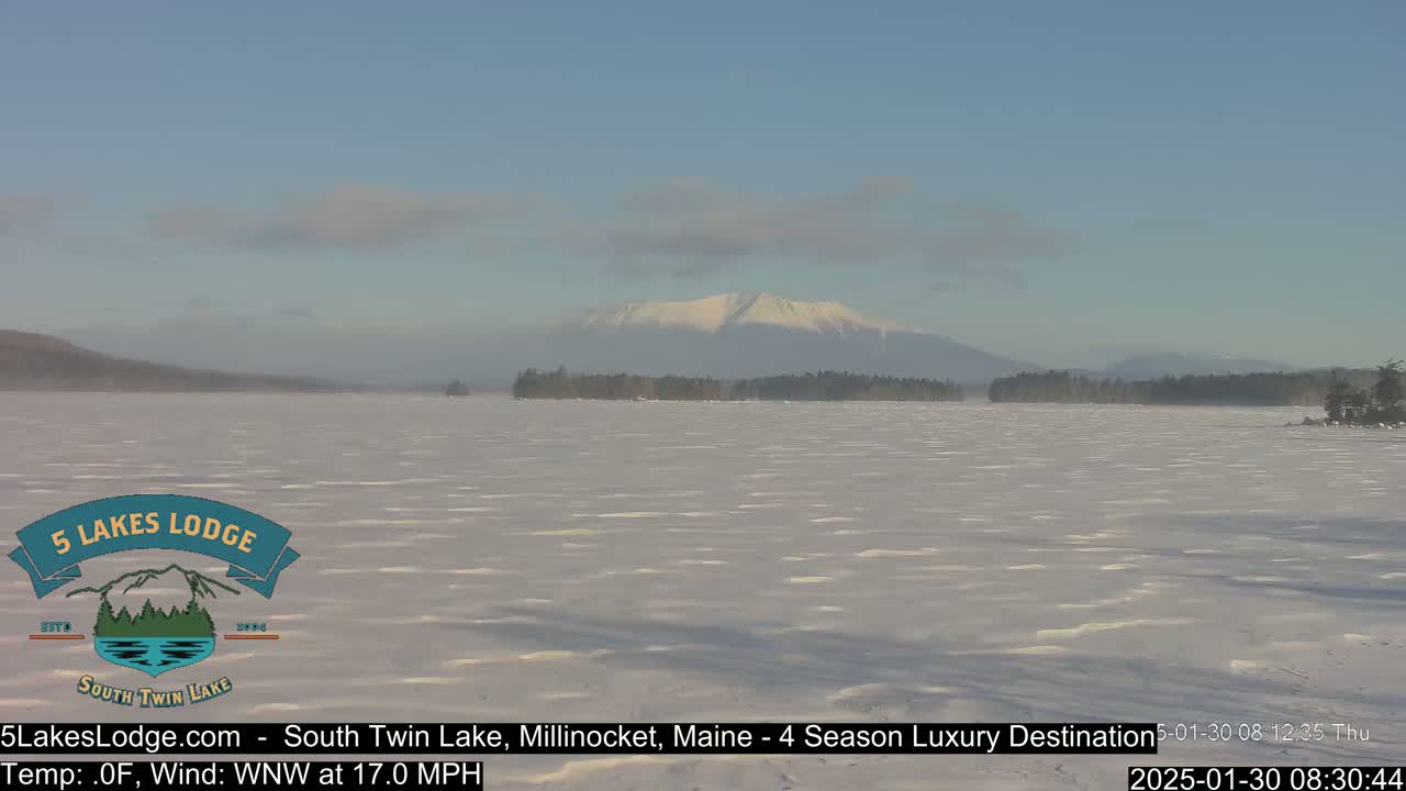 A snow-covered lake stretches to a distant, snow-capped mountain range under a clear, bright sky; a brisk wind is blowing.