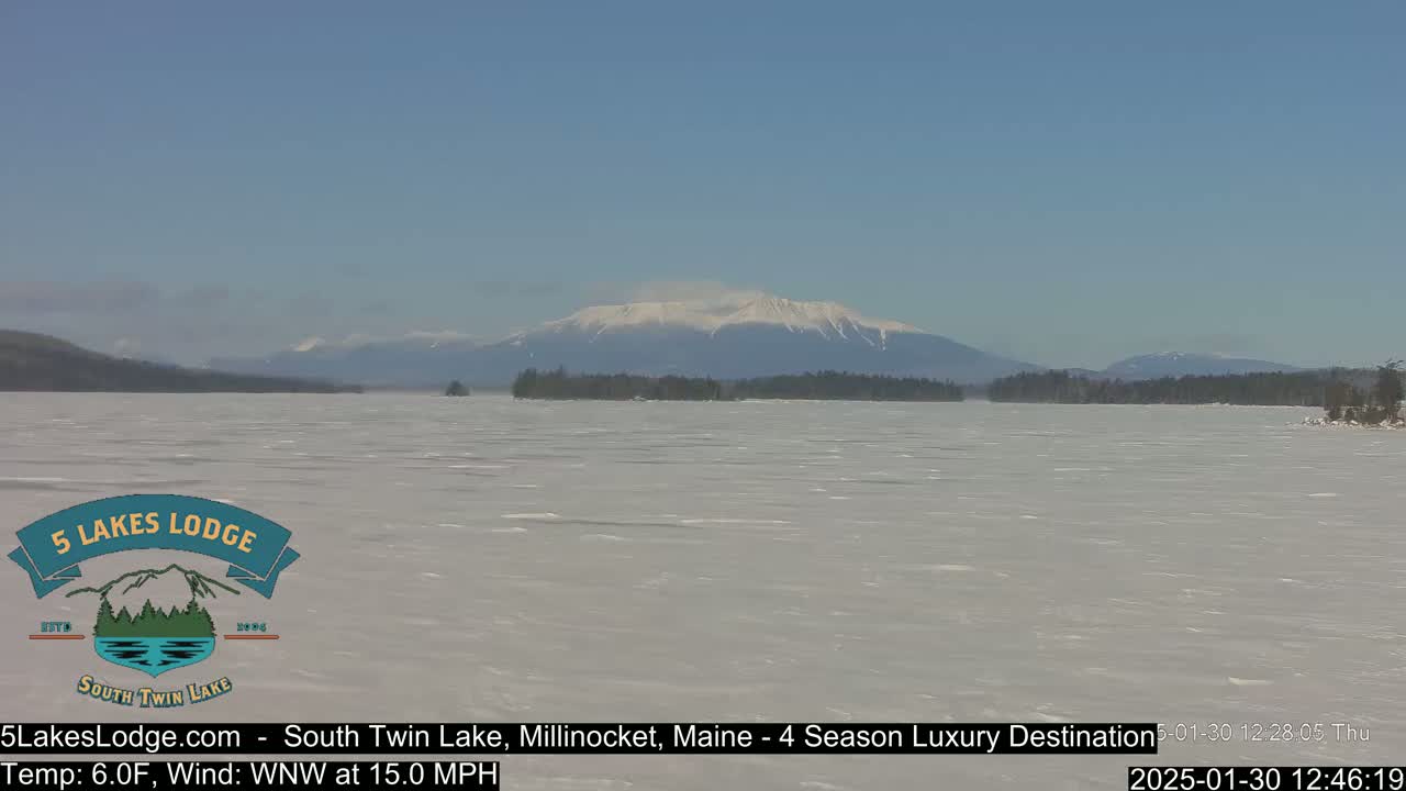 A snow-covered lake stretches to a mountain range with a snow-capped peak under a clear blue sky.
