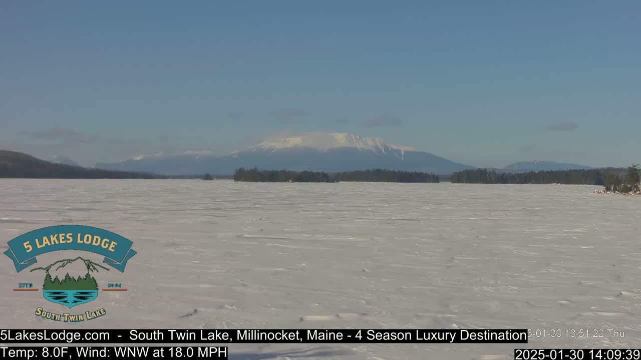 A frozen lake stretches towards snow-capped mountains under a clear blue sky, with a brisk wind blowing.