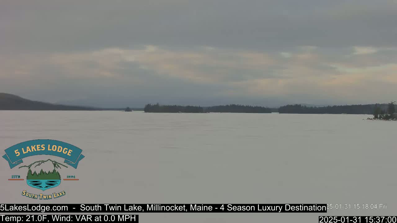 A snow-covered lake stretches to a distant treeline under a cloudy sky.