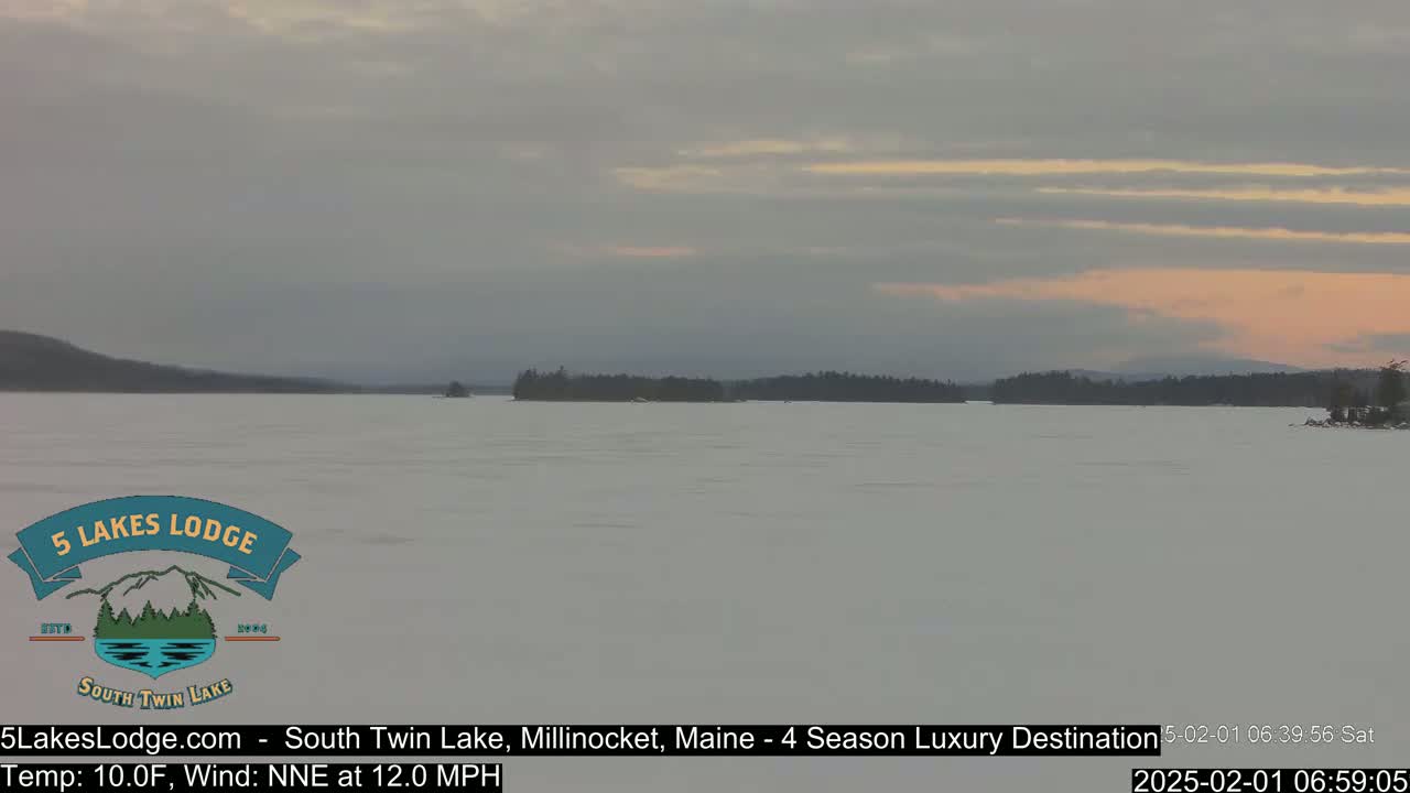 A frozen lake stretches to a snow-covered shoreline under a pale, pinkish sky.